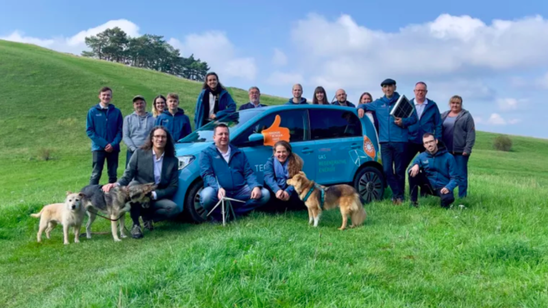 Auf einer grünen Wiese stehen die Mitarbeiter:innen der Teckwerke Bürgerenergie eG um ein blaues Auto in einer Hügellandschaft.