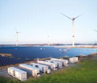 Container mit Batteriespeichern im Vordergrund von einem Solarpark. Im Hintergrund: Windenergieanlagen.