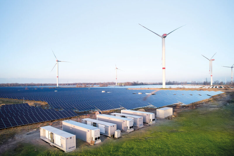 Container mit Batteriespeichern im Vordergrund von einem Solarpark. Im Hintergrund: Windenergieanlagen.