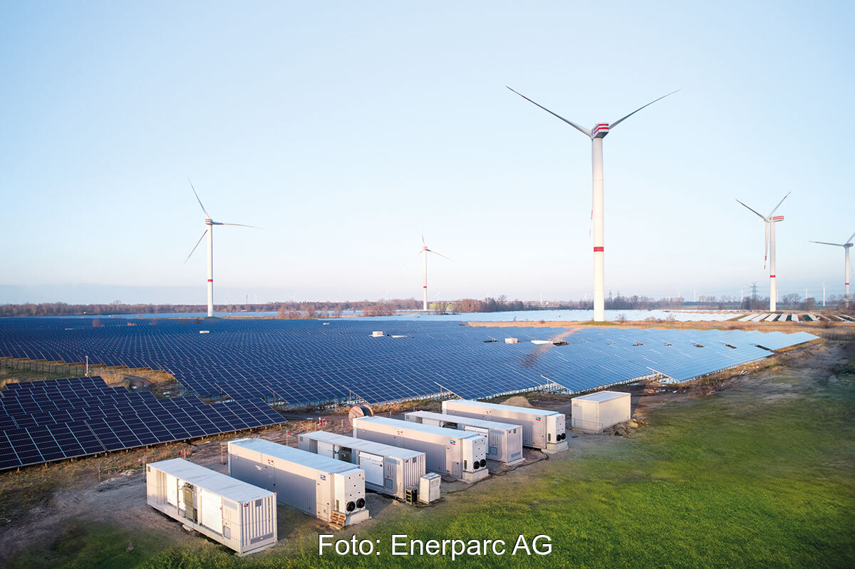 Container mit Batteriespeichern im Vordergrund von einem Solarpark. Im Hintergrund: Windenergieanlagen.