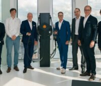 Sieben Männer stehen in einem hellen Büro neben einer schwarzen Ladesäule für Elektrofahrzeuge und blicken in die Kamera. Zwei Personen halten Ladekabel in der Hand. Im Hintergrund sind bodentiefe Fenster mit Blick auf eine Stadt zu sehen.