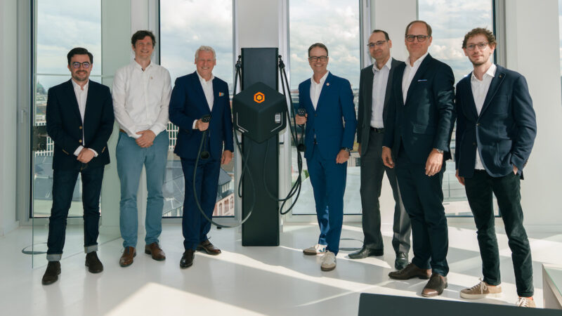 Sieben Männer stehen in einem hellen Büro neben einer schwarzen Ladesäule für Elektrofahrzeuge und blicken in die Kamera. Zwei Personen halten Ladekabel in der Hand. Im Hintergrund sind bodentiefe Fenster mit Blick auf eine Stadt zu sehen.