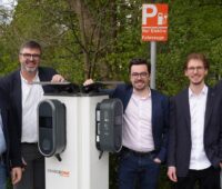 Auf dem Foto sind fünf dunkelhaarige Männer mit dunklen Sakkos und hellen Hemden zu sehen. Sie stehen um eine hellgraue Ladesaäule, zwei links, drei rechts. Im Hintergrund dunkelgrüne Sträucher.