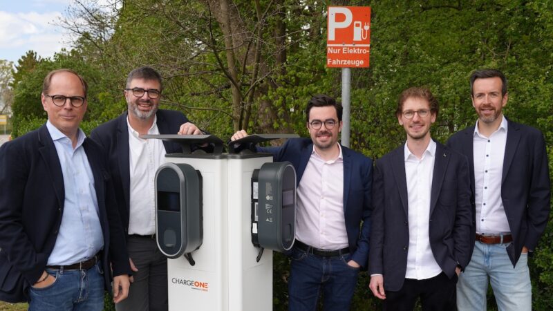 Auf dem Foto sind fünf dunkelhaarige Männer mit dunklen Sakkos und hellen Hemden zu sehen. Sie stehen um eine hellgraue Ladesaäule, zwei links, drei rechts. Im Hintergrund dunkelgrüne Sträucher.