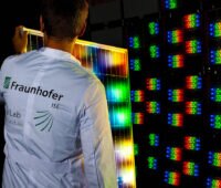 Mitarbeiter des Fraunhofer ISE hält ein PV-Modul vor einen multispektralen Sonnensimulator mit roten, grünen und blauen LEDs.