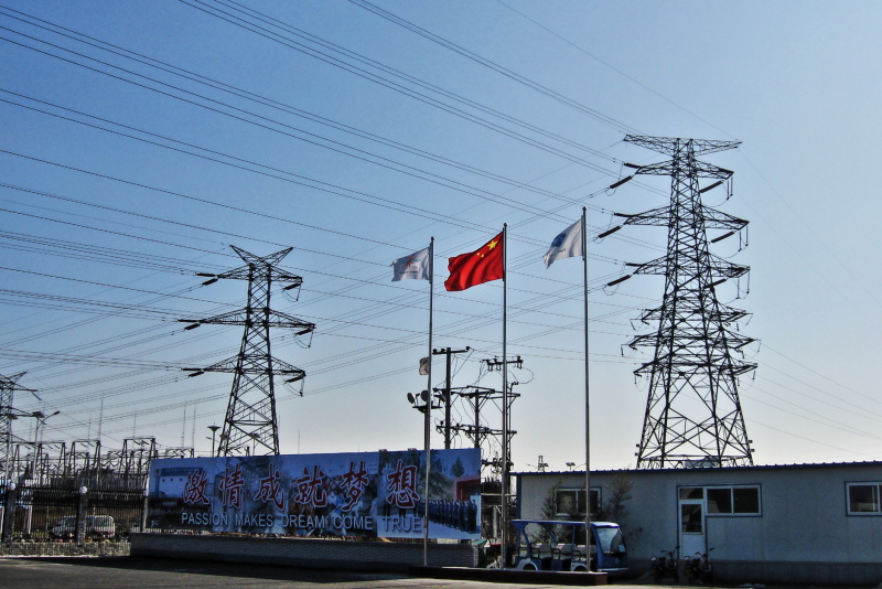 Strommasten und Leitungen vor blauem Himmel. Mittig eine wehende chinesische Fahne.