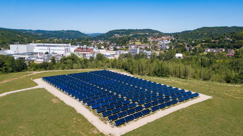 Eine solarthermische Kollektoranlage auf grüner Wiese vor einer Fabrik und Hügeln im Hintergrund.