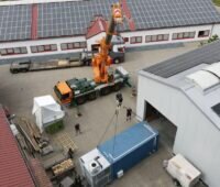 Luftaufnahme eines Containers, der mit einem Kran aus einer Halle mit PV-Modulen heraus entladen wird.