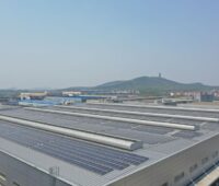 Reihen mit Solarmodulen auf einem großen flachen Industriedach, im Hintergrund Berge und ein Turm.