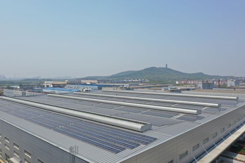 Reihen mit Solarmodulen auf einem großen flachen Industriedach, im Hintergrund Berge und ein Turm.