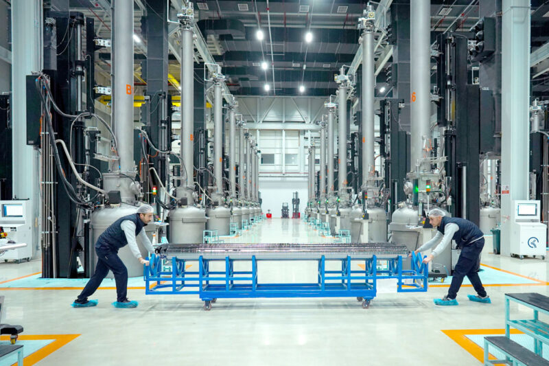 Fabrik für Silizium-Ingots. Mitarbeiter schieben in Reinraum-Montur einen Wagen mit Einkristallen. Im Hintergrund: eine Reihe von Siemens-Öfen.