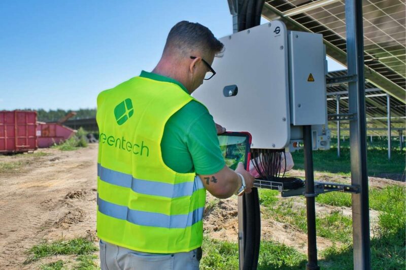 Ein Mann in gelber Warnweste mit Aufschrift "greetech" fotografiert mit einem Tablet die Kabelstränge an einem Wechselrichter auf einer Freiflächen Photovoltaik Anlage.