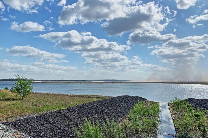 Blick über einen künstlichen Binnensee mit Kohle-Kühltürmen am Horizont sowie Windkraftanlagen