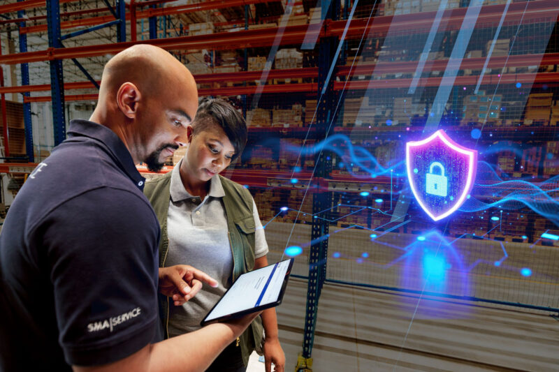 Symbolbild für Cybersicherheit: Mann und Frau mit Tablet-Computer in einem Lagerraum - Lichteffekte projezieren ein Schloss inn den Raum.