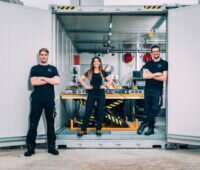 Zwei Männer und eine Frau in schwarz an einem Container mit technischem Equipment.