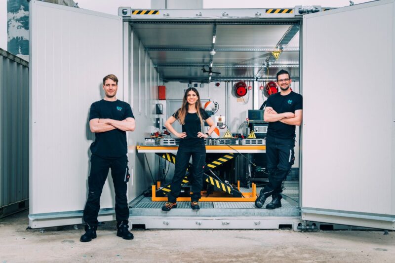 Zwei Männer und eine Frau in schwarz an einem Container mit technischem Equipment.