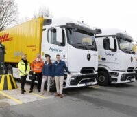 Vier Personen stehen neben einer weißen eActros-Zugmaschine plus einem gelbem DHL-Anhänger. Schräg dahinter zwei weitere E-LKW.