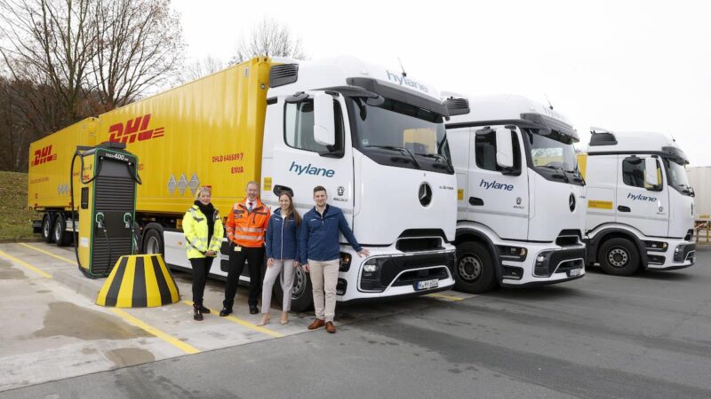 Vier Personen stehen neben einer weißen eActros-Zugmaschine plus einem gelbem DHL-Anhänger. Schräg dahinter zwei weitere E-LKW.