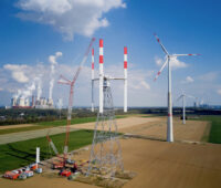 Aufbau einer Windkraftanlage mit vertikal angeordneten Rotoblättern auf dem Windtestfeld - rechts weitere "normale" Windkraftanlagen und links im Hintergrund ein Kohlekraftwerk