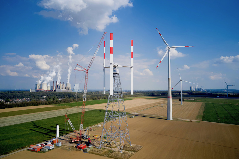 Aufbau einer Windkraftanlage mit vertikal angeordneten Rotoblättern auf dem Windtestfeld - rechts weitere "normale" Windkraftanlagen und links im Hintergrund ein Kohlekraftwerk