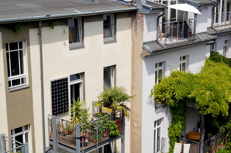 Eine Hausfassade, links vor im Bild ein Balkon mit Pflanzen und einem Photovoltaikmodul an der Wand, einer Balkon-Solaranlage.