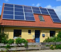 Foto eines Ein-familienhauses mit Garten und Bäumen. Auf dem Dach befindet sich eine PV-Anlage.