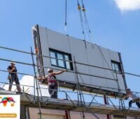 Ein Fassadenelement mit Fensteröffnung wird an einem Kran zu einem Haus heruntergelassen - Serielle Sanierung.