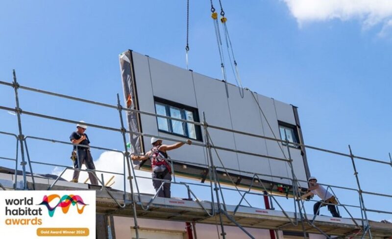 Ein Fassadenelement mit Fensteröffnung wird an einem Kran zu einem Haus heruntergelassen - Serielle Sanierung.