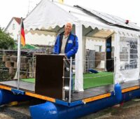 Der Bastler Wille Riedel steht auf einem Floß an Land mit einem Zelt überspannt, auf dem Solarmodule installiert sind