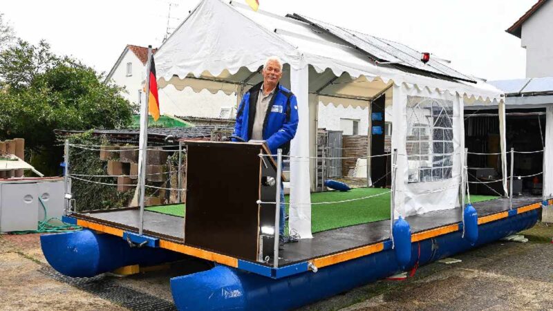 Der Bastler Wille Riedel steht auf einem Floß an Land mit einem Zelt überspannt, auf dem Solarmodule installiert sind