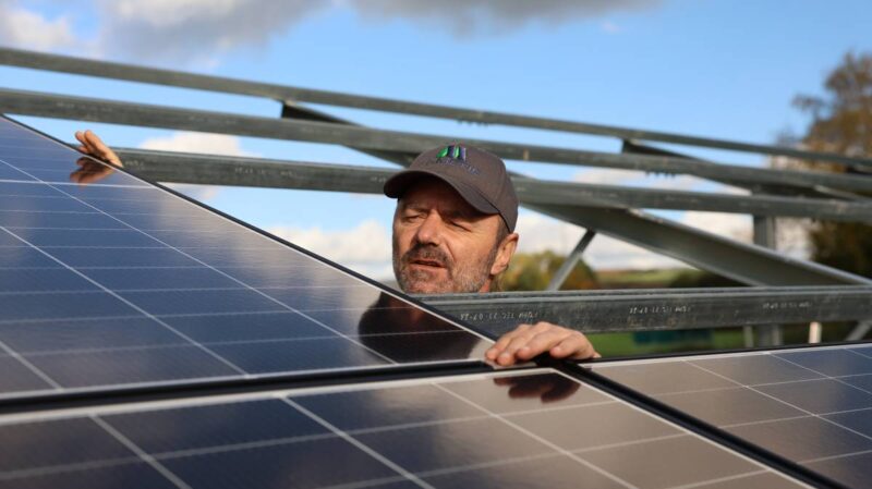 Installateuer lugt zwischen Solarmodulen hervor, die auf Unterkonstruktion ruhen.