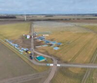 Das Foto zeigt das zukünftige Agri-PV-Gelände aus der Vogelperspektive von schräg vorn, mit Horizont und einem bewölkten Himmel: Auf den Ackerflächen entstehen die Unterkonstruktionen für die Photovoltaik-Module. Baumaterial lagert im Vordergrund der Baustelle, Fahrzeuge stehen auf dem Weg zum Gelände.