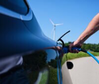 Nahaufnahme eines Arms mit Ladekabel zeigt, wie eine Person ein Ladekabel an ein E-Auto steckt. Im Hintergrund ein Windrad.
