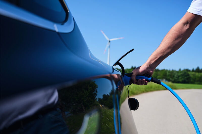 Nahaufnahme eines Arms mit Ladekabel zeigt, wie eine Person ein Ladekabel an ein E-Auto steckt. Im Hintergrund ein Windrad.