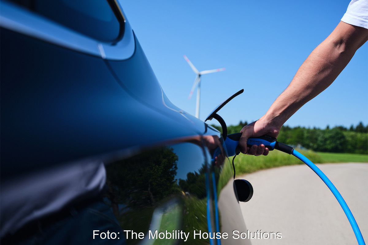 Nahaufnahme eines Arms mit Ladekabel zeigt, wie eine Person ein Ladekabel an ein E-Auto steckt. Im Hintergrund ein Windrad.