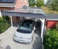 Graues Elektroauto unter einem mit Solarmodulen belegten Carport vor einem Backsteinhaus mit weiterer Photovoltaikanlage.