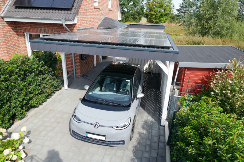 Graues Elektroauto unter einem mit Solarmodulen belegten Carport vor einem Backsteinhaus mit weiterer Photovoltaikanlage.