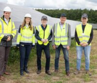 Fünf Personen stehen bei einer Einweihung in neongelben Warnwesten vor einem Solarmodul in einem Solarpark.