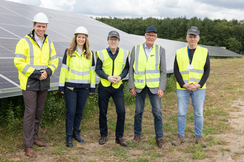 Fünf Personen stehen bei einer Einweihung in neongelben Warnwesten vor einem Solarmodul in einem Solarpark.