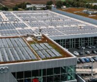 Aufsicht auf das Dach einer Gewerbehalle mit Audi-Logo, den vier Ringen an der Seite; auf der vorderen Dachfläche ist eine PV-Anlage zu sehen.