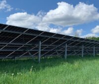 Auf dem Foto ist eine Reihe einer Photovoltaik-Freiflächenanlage auf Rasen zu sehen, aufgenommen von der Rückseite, so dass das Gestänge zu sehen ist.