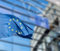 Eine EU-Flagge weht vor einem modernen Glasgebäude in Brüssel, das im Hintergrund unscharf zu sehen ist.