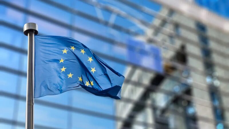 Eine EU-Flagge weht vor einem modernen Glasgebäude in Brüssel, das im Hintergrund unscharf zu sehen ist.