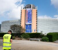 Am Sitz der EU-Kommission in Brüssel hängt ein Plakat mit der Aufschrift EU NExt Generation, im Vordergrund in Arbeiter in Sicherweste mit dem Rücken zum Betrachter auf den Boden schauend.