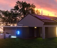 Haus mit Photovoltaikanlage vor rotem Abendhimmel. Im Vordergrund lädt ein Elektroauto