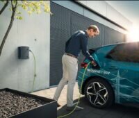 Vor der Fassade eines Einfamilienhauses, an der Garage, steckt ein Mann das Ladekabel in sein Elektrofahrzeug.