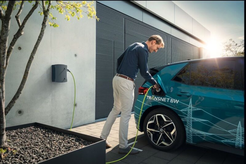 Vor der Fassade eines Einfamilienhauses, an der Garage, steckt ein Mann das Ladekabel in sein Elektrofahrzeug.