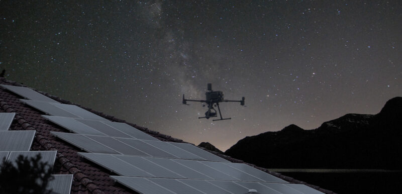Drohne schwebt über Dach-PV-Anlage vor Sternenhimmel - Messung mit Elektrolumineszenz-Verfahren.