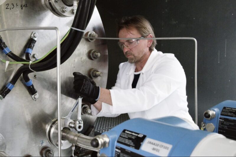 Ein Mann im weißen Kittel und mit Schutzbrille arbeitet mit einem Werkzeug an einem Elektrolyse-Block.