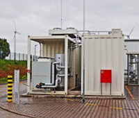 Zu sehen ist ein weißes Metallcontaiber. Darin befindet sich ein Elektrolyseur zur Wasserstoffproduktion der Stadtwerke Mainz und Linde. Im Hintergrund stehen Windkraftanlagen